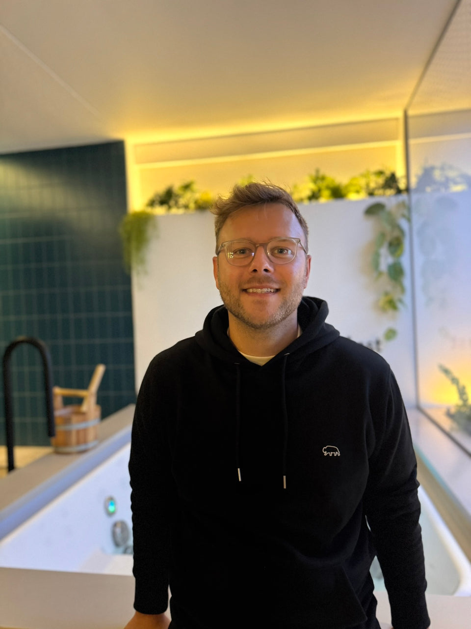 Man wearing a black breitung hoodie with a bison ouline logo, standing in a modern indoor setting.