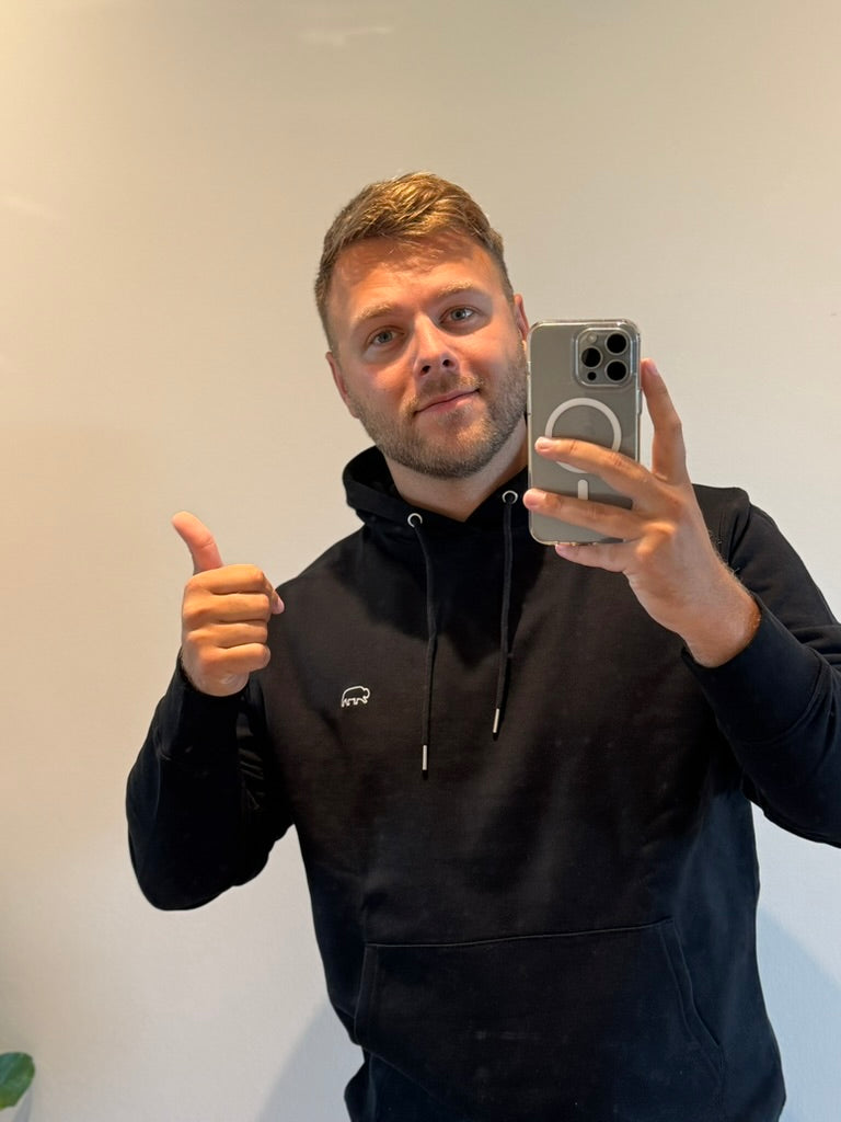 Man taking a mirror selfie wearing a black breitung hoodie with a logo, giving a thumbs-up.