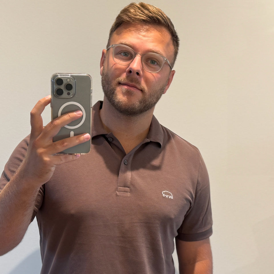 Man wearing a brown Breitung polo shirt with a logo, holding a phone with a gray case.